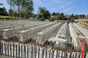 Zdjęcie przedstawia zaawansowany etap budowy mostu, na którym widać zamontowane prefabrykowane belki betonowe. Belki te są ustawione równolegle, tworząc solidną podstawę dla przyszłej nawierzchni mostu. Widoczne są liczne elementy zbrojeniowe wystające z górnej części belek, które będą służyć do połączenia kolejnych warstw konstrukcji mostu. Na pierwszym planie zdjęcia widoczne są pręty zbrojeniowe, wystające pionowo z betonowej konstrukcji. Te elementy są kluczowe dla dalszego wzmocnienia struktury mostu. W tle, po prawej stronie, widać stosy prętów zbrojeniowych, które czekają na dalsze wykorzystanie. Otoczenie budowy jest zielone, z drzewami po lewej stronie i w tle, co pokazuje, że most powstaje w malowniczym, naturalnym krajobrazie. Na horyzoncie widać kilka domów, co ukazuje, że budowa odbywa się w pobliżu obszarów zamieszkałych. Jasne, słoneczne niebo świadczy o dobrych warunkach pogodowych w dniu wykonania zdjęcia. Cała scena oddaje dynamikę pracy na placu budowy, gdzie widać postępy w realizacji projektu mostu.