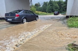 Samochody jadą przez zalaną wodą drogę pod wiaduktem. Wysoki poziom wody i błotnisty kolor wskazują na poważne podtopienie po intensywnych opadach deszczu. W tle widać zieleń i fragment drogi.