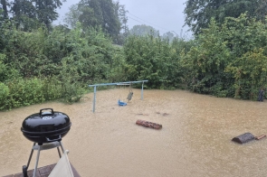  Zalany ogród z huśtawką, grill węglowy i unoszące się na wodzie przedmioty. Woda sięga ponad trawnik, otaczając gęste krzewy i drzewa. Intensywne opady deszczu spowodowały lokalne podtopienia.