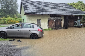 Samochód osobowy stoi na zalanym podwórku obok budynku z garażem. Woda sięga niemal do połowy kół auta. W tle widać znaki drogowe i bujną roślinność. 