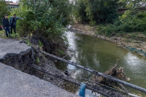 Dwóch mężczyzn, w tym Rajmund Dedio, Burmistrz Skoczowa stoją na skraju drogi, z której znaczna część została zerwana przez wodę. Obaj wskazują na erozję i zniszczenia wzdłuż brzegu rzeki, gdzie widać zwalone drzewa i zniszczone fragmenty infrastruktury. Zdjęcie dokumentuje skutki powodzi i pokazuje skalę zniszczeń spowodowanych przez żywioł.