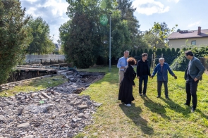 Pięć osób, w tym Janina Żagan, Starosta stoją na trawniku, rozmawiają ze sobą. W tle widoczne są drzewa, domy i uszkodzony teren z rozrzuconymi kawałkami nawierzchni, co pokazuje, że jest to miejsce dotknięte przez powódź. Jeden z mężczyzn pokazuje ręką obszar zniszczeń.