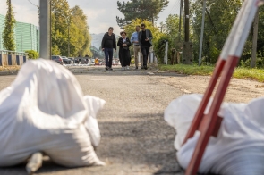  Cztery osoby, w tym Janina Żagan Starosta Cieszyński oraz Marek Wójcik, Wojewoda Śląski spacerują wzdłuż drogi w Skoczowie, na której znajdują się worki z piaskiem, zabezpieczające przed skutkami powodzi. W tle widoczne są samochody i elementy zabezpieczeń, a także zielona roślinność po obu stronach drogi. Osoby te prowadzą rozmowę, omawiając sytuację po powodzi. 