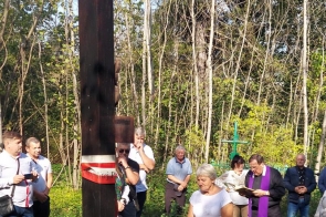 Uczestnicy uroczystości składają wieniec pod drewnianym krzyżem w leśnej scenerii. Obecny jest ksiądz z fioletową stułą, modlący się. W tle zgromadzeni ludzie w zadumie. 