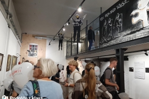 Na zdjęciu widoczni są uczniowie zwiedzający muzeum, w którym znajdują się prace artystyczne Banksy'ego. Na ścianach znajdują się charakterystyczne dla Banksy'ego murale, w tym postacie aniołów oraz inne elementy street artu. Grupa ludzi ogląda ekspozycję, w rękach trzymają balony z logo "Muzeum Banksy'ego". W tle widać także balustradę. 