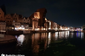 Zdjęcie ukazuje nocną panoramę Gdańska z widokiem na Żuraw nad rzeką Motławą. Zabytkowe kamienice i oświetlone nabrzeże odbijają się w wodzie, tworząc malowniczy krajobraz miasta po zmroku. Charakterystyczna sylwetka Żurawia, jednego z najbardziej rozpoznawalnych symboli Gdańska, jest centralnym punktem kompozycji. W dolnym lewym rogu zdjęcia znajduje się logo „Podróże z klasą”. 