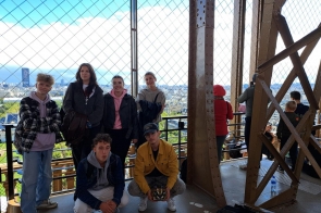  Grupa młodych osób stoi na jednym z poziomów wieży Tower of London, z której rozpościera się panoramiczny widok na miasto. W tle widać charakterystyczne londyńskie budynki oraz niebo pokryte lekkimi chmurami. Młodzież, ubrana w wygodne, codzienne stroje, z uśmiechami na twarzach pozuje do zdjęcia, niektóre osoby stoją, inne kucają. Atmosfera na zdjęciu jest swobodna, a otaczająca siatka zabezpieczająca podkreśla wysokość i monumentalność budowli.