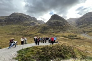  Na zdjęciu widoczna jest grupa osób pozujących na tle majestatycznych gór w Dolinie Glencoe w Szkocji. W oddali dominują trzy charakterystyczne szczyty, zwane „Trzema Siostrami”, które majestatycznie wznoszą się nad otaczającą je doliną. Górskie krajobrazy porośnięte trawą i wrzosami tworzą idylliczną scenerię, a chmury przysłaniają niebo, nadając miejscu tajemniczy charakter. Grupa turystów stoi na ścieżce prowadzącej w głąb gór, niektóre osoby pozują w dynamicznych pozach, co dodaje zdjęciu energii i radości z odkrywania przyrody.