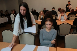 Kobieta z długimi, ciemnymi włosami rozdaje testy uczestnikom Olimpiady. Podchodzi do stołu. Obok niej siedzi młoda dziewczyna w szarej bluzce, która wygląda na zamyśloną. W tle widać innych uczestników, siedzących przy stołach, zajęci są rozmową. Sala jest przestronna, w tle znajduje się stolik z napojami i przekąskami. 