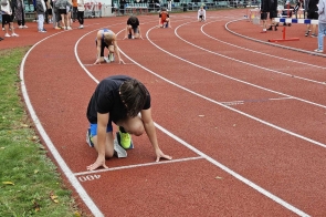 Zdjęcie przedstawia grupę młodych sportowców przygotowujących się do startu w biegu na tartanowej bieżni stadionu. Uczestnicy są ustawieni w blokach startowych, a atmosfera koncentruje się na nadchodzącym wysiłku. W tle widać innych zawodników i osoby na trybunach.