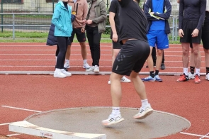 Osoba w czarnym stroju sportowym wykonuje rzut kulą na stadionie lekkoatletycznym. W tle grupa młodych ludzi obserwuje, część z nich uśmiecha się. Na drugim planie widoczne są budynki oraz ogrodzenie. Atmosfera skupienia i sportowej rywalizacji.