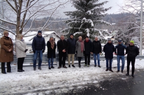 Grupa kilkunastu osób stoi w rzędzie na zaśnieżonej drodze, ubrana w zimowe płaszcze i kurtki. W tle widoczne są drzewa pokryte śniegiem oraz dachy domów w zimowej scenerii.