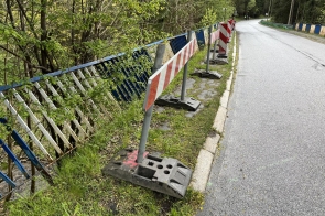 Na asfaltowej drodze widać metalowe barierki, częściowo zniszczone, otoczone bujną zielenią. Na chodniku ustawiono przenośne znaki ostrzegawcze z biało-czerwonymi pasami.