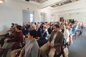 Na zdjęciu widoczna jest grupa ludzi siedzących w rzędach na sali konferencyjnej. Pomieszczenie jest jasne, z dużymi oknami wpuszczającymi naturalne światło. Uczestnicy są elegancko ubrani, skupieni na słuchaniu. W tle widać wieszaki z płaszczami i otwarte drzwi do innego pomieszczenia.