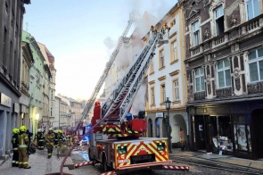 Na zdjęciu widoczna akcja strażacka w centrum miasta Cieszyna (ul. Głęboka). Kilka wozów strażackich, drabiny skierowane na płonący budynek, z którego unosi się biały dym. Strażacy w żółtych kaskach i mundurach pracują na chodniku oraz przy pojazdach. W tle rząd zabytkowych kamienic.