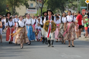 Barwny korowód uczestników Dożynek Powiatowo-Gminnych w Brennej. Grupa osób, ubrana w tradycyjne stroje ludowe, maszeruje ulicą, niosąc ozdobny wieniec dożynkowy z kwiatami słonecznika i biało-czerwoną wstążką. Kobiety mają na sobie kolorowe spódnice i bluzki z haftowanymi wzorami, a mężczyźni są ubrani w ludowe kamizelki i kapelusze. W tle widać innych uczestników korowodu oraz widownię, która obserwuje to wydarzenie. Na pierwszym planie widać również tabliczkę z napisem „Mała Brenna”, wskazującą na udział delegacji z tej miejscowości. Atmosfera zdjęcia jest radosna i pełna życia, co podkreśla tradycyjny charakter uroczystości.