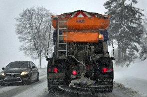 Zaśnieżona droga, którą odśnieża pługopiaskarka jadąca z prawej strony, na drugim pasie jedzie czarne auto. W tle widać zaśnieżone drzewa, pobocze jest zasypane śniegiem, pada śnieg.