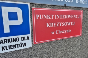 Zdjęcie przedstawia dwa znaki na ścianie budynku. Jeden to niebieski znak parkingowy z literą „P” i napisem „PARKING DLA KLIENTÓW”. Drugi to czerwona tablica z białym napisem „PUNKT INTERWENCJI KRYZYSOWEJ w Cieszynie”, oznaczająca miejsce pomocy.