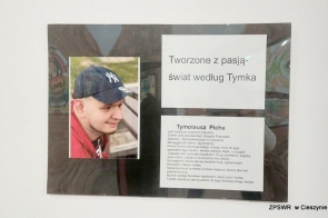 Tablica prezentuje portret młodego mężczyzny w czapce z daszkiem oraz tekst zatytułowany „Tworzone z pasją – świat według Tymka”. Poniżej umieszczono opis opowiadający o osobie, jej życiu i twórczości. W tle widać delikatne motywy artystyczne, co podkreśla kreatywność autora. Całość sprawia wrażenie hołdu złożonego pasji i talentowi.