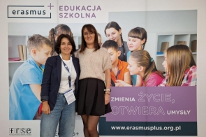 Karolina Grabowska oraz Alicja Wach stoją na tle baneru promującego program Erasmus+ „Edukacja Szkolna”. Na banerze znajduje się napis: „Zmienia życie, otwiera umysły” oraz adres strony internetowej: www.erasmusplus.org.pl.