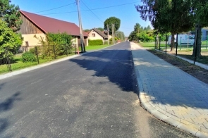  Zdjęcie przedstawia nowo wyremontowaną ulicę Wiejską w Drogomyślu. Widoczna jest świeżo położona asfaltowa nawierzchnia oraz nowy chodnik po prawej stronie. Ulica otoczona jest domami jednorodzinnymi, ogrodzeniami i zielenią.