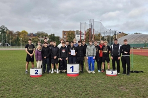 Grupa młodych sportowców w sportowych strojach stoi na murawie boiska obok podium z cyframi 1, 2 i 3. W centrum znajduje się kobieta, a jeden z uczestników trzyma dyplom. W tle widać elementy stadionu lekkoatletycznego, budynki oraz pochmurne niebo.