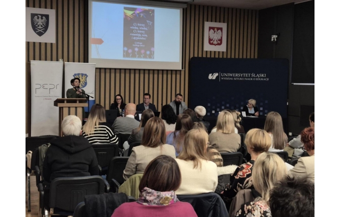 Zdjęcie przedstawia uczestników konferencji zgromadzonych w sali wykładowej Wydziału Sztuki i Nauk o Edukacji Uniwersytetu Śląskiego w Cieszynie. Przy mównicy przemawia Magdalena Bosowska, natomiast za stołem prezydialnym siedzą inni specjaliści. Na ekranie za nimi wyświetlony jest slajd, który zawiera cytat: „Ci, którzy wiedzą, wiedzą. Ci, którzy rozumieją, uczą”. Obok wyświetla się logo Uniwersytetu Śląskiego oraz Powiatowego Centrum Pomocy Rodzinie w Cieszynie. Konferencja dotyczyła tematyki przemocy wobec dzieci, a jej celem była wymiana wiedzy i doświadczeń w walce z tym problemem.