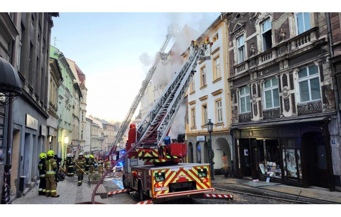 Na zdjęciu widoczna akcja strażacka w centrum miasta Cieszyna (ul. Głęboka). Kilka wozów strażackich, drabiny skierowane na płonący budynek, z którego unosi się biały dym. Strażacy w żółtych kaskach i mundurach pracują na chodniku oraz przy pojazdach. W tle rząd zabytkowych kamienic.