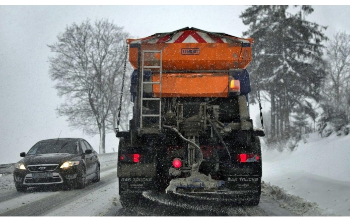 Zaśnieżona droga, którą odśnieża pługopiaskarka jadąca z prawej strony, na drugim pasie jedzie czarne auto. W tle widać zaśnieżone drzewa, pobocze jest zasypane śniegiem, pada śnieg.