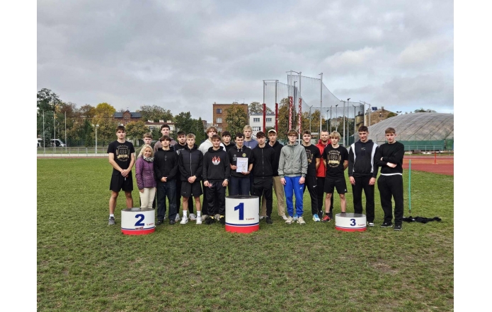 Grupa młodych sportowców w sportowych strojach stoi na murawie boiska obok podium z cyframi 1, 2 i 3. W centrum znajduje się kobieta, a jeden z uczestników trzyma dyplom. W tle widać elementy stadionu lekkoatletycznego, budynki oraz pochmurne niebo.