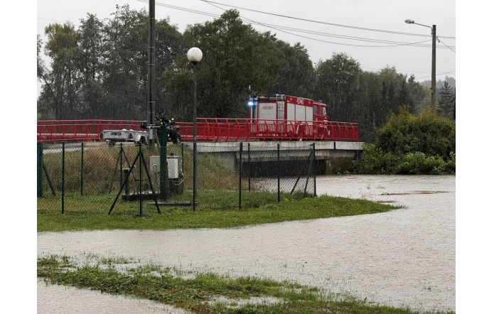 Straż pożarna stoi na moście podczas intensywnego deszczu. Woda z pobliskiego terenu zaczyna się rozlewać, co pokazuje zagrożenie powodziowe. W tle widać drzewa, a na pierwszym planie zielony teren ogrodzony siatką.