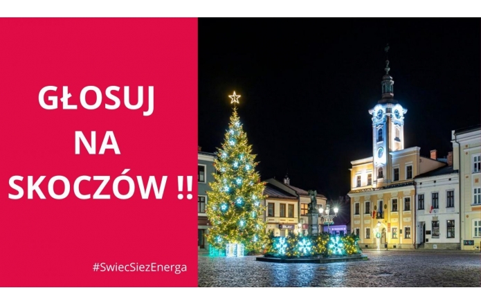 Na zdjęciu widoczny jest rynek w Skoczowie nocą, pięknie oświetlony świątecznymi dekoracjami. Centralnie stoi udekorowana choinka z gwiazdą na czubku, otoczona jasnymi, migoczącymi ozdobami w kształcie prezentów. W tle ratusz z podświetloną wieżą zegarową, dodający uroku tej zimowej scenerii.