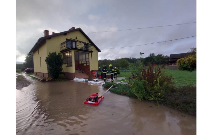 Strażacy wypompowują wodę z zalanej posesji przy domu jednorodzinnym. Woda sięga schodów wejściowych. Na pierwszym planie widoczna jest pompa.