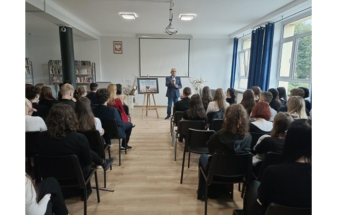 Na zdjęciu widać grupę uczniów siedzących w sali, którzy uczestniczą w spotkaniu z okazji Światowego Dnia Turystyki. Na środku, przed grupą, stoi prelegent w garniturze, który przemawia do zebranych. W tle widać ekran projekcyjny i tablicę informacyjną z napisem "Światowy Dzień Turystyki". Sala jest jasna, z dużymi oknami i regałami z książkami, co nadaje miejscu atmosferę nauki i skupienia.