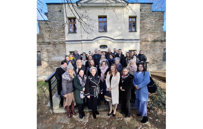 Grupa ludzi pozuje na tle Dworu Kossaków o jasnej fasadzie z widocznymi oknami i fragmentami ceglanej ściany. Wszyscy ubrani w jesienne płaszcze, uśmiechają się. Scena odbywa się w słoneczny dzień, w otoczeniu suchych liści i bezlistnych gałęzi. Atmosfera przyjazna i formalna.