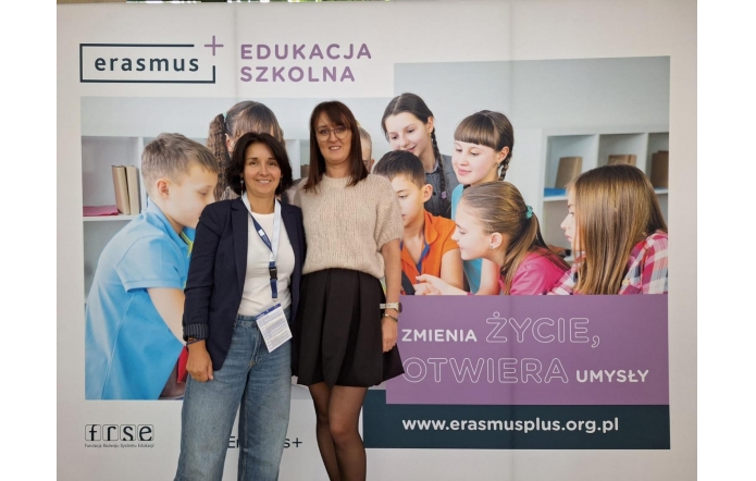 Karolina Grabowska oraz Alicja Wach stoją na tle baneru promującego program Erasmus+ „Edukacja Szkolna”. Na banerze znajduje się napis: „Zmienia życie, otwiera umysły” oraz adres strony internetowej: www.erasmusplus.org.pl.