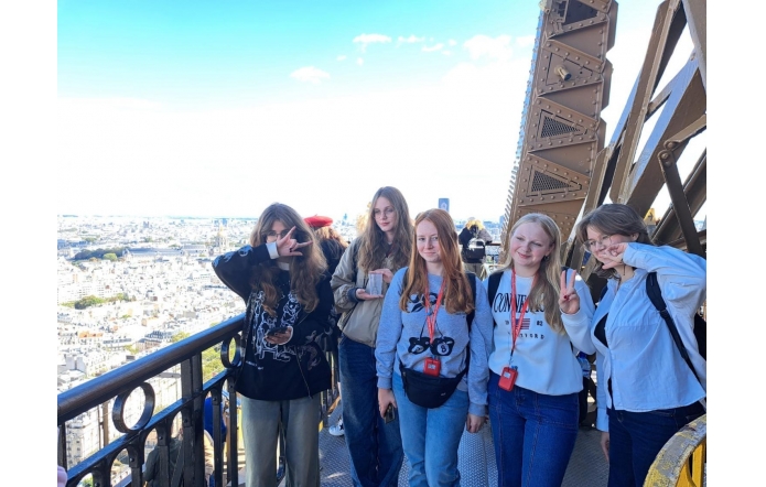 Grupa uczennic stoi na jednej z platform widokowych Tower of London. W tle rozciąga się panorama miasta z charakterystycznymi budynkami i błękitnym niebem. Dziewczęta uśmiechają się, pozując w luźnej atmosferze, niektóre wykonują gesty rękoma, dodając zdjęciu nieformalnego, przyjacielskiego charakteru. Ich identyfikatory na smyczach wskazują, że uczestniczą w zorganizowanej wycieczce.