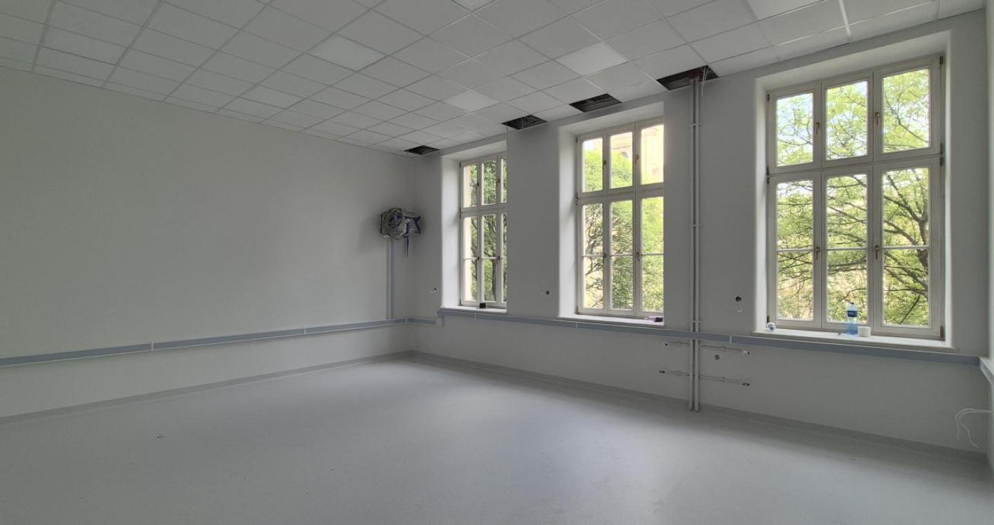 Wyremontowane pomieszczenie z jasnymi ścianami, dużymi oknami i nową podłogą. Przy ścianach widoczne są elementy instalacji, a w suficie znajdują się częściowo otwarte panele.