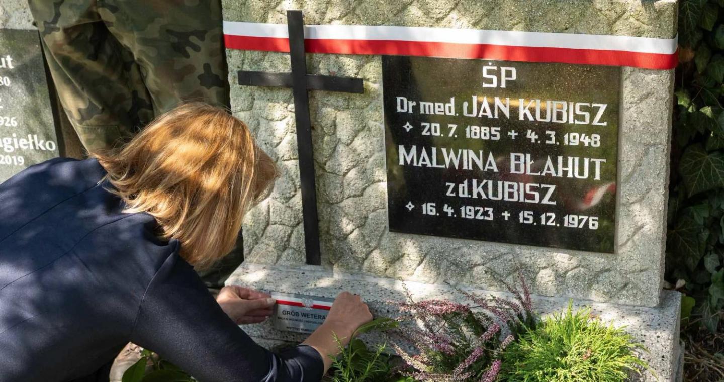 Kobieta przy grobie ozdobionym biało-czerwoną wstęgą umieszcza oznaczenie na płycie nagrobnej. Wokół widoczne są rośliny oraz kwiaty.