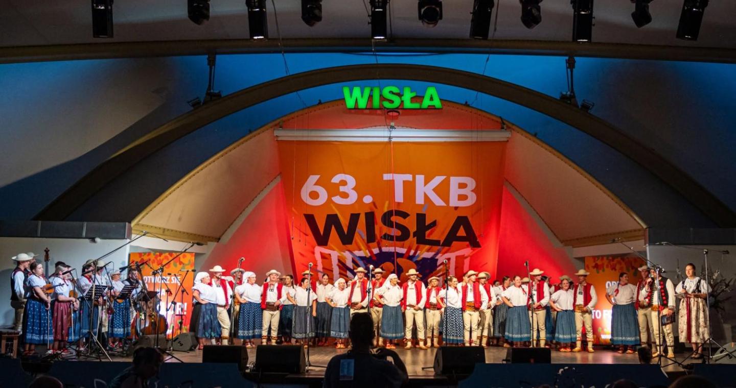 Grupa osób w strojach regionalnych stoi w rzędzie na scenie. Po lewej stronie widoczni są muzycy z instrumentami, a po prawej dwie kobiety stoją przy mikrofonach. W tle znajduje się napis „63. TKB Wisła”. Oświetlenie sali jest ciemne, a światło sceniczne pada głównie na stojące osoby.