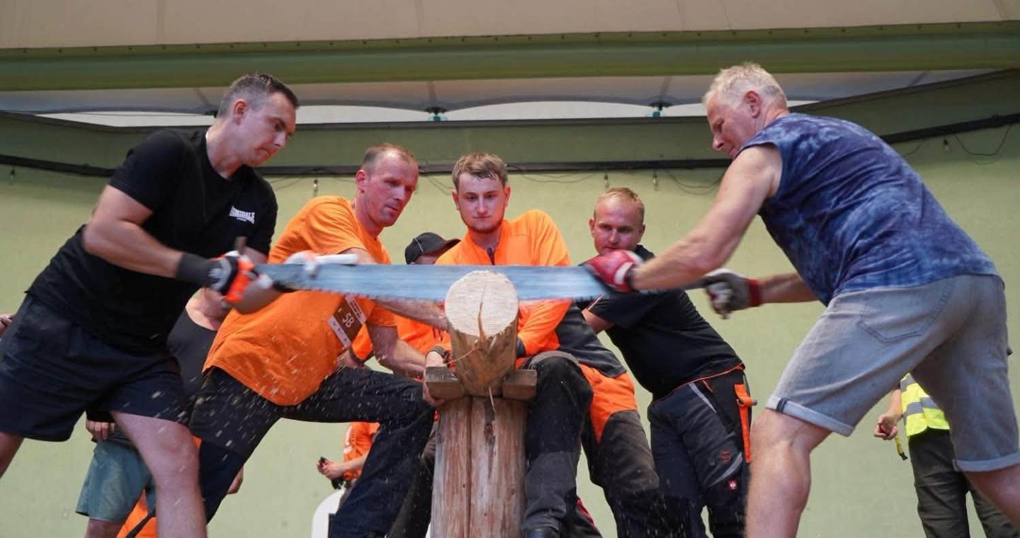Pięciu mężczyzn przecina drewnianą belkę dużą piłą ręczną. Trzech z nich ma pomarańczowe koszulki, a pozostali czarne i granatowe ubrania. W tle stoją kolejne osoby.