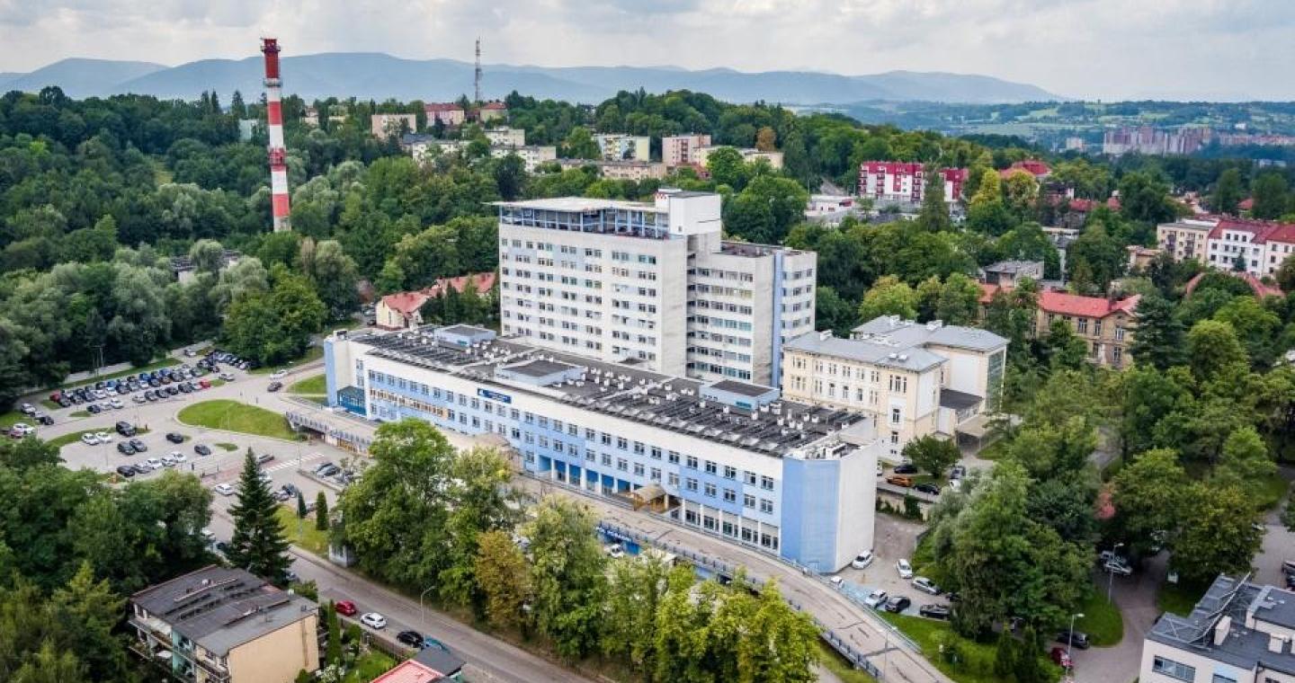Rozległy kompleks budynków otoczony zielenią, widziany z lotu ptaka. Wokół znajdują się drogi, parkingi i zabudowa miejska, a w tle widoczne są wzgórza i góry.