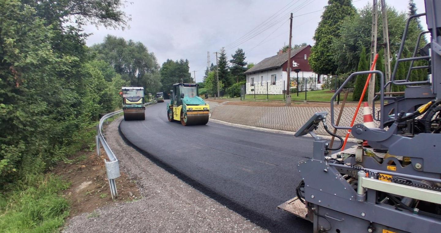 Maszyny drogowe układają i zagęszczają nową warstwę asfaltu na jezdni. Wzdłuż drogi widoczne są bariery ochronne, chodnik oraz zabudowania otoczone zielenią.