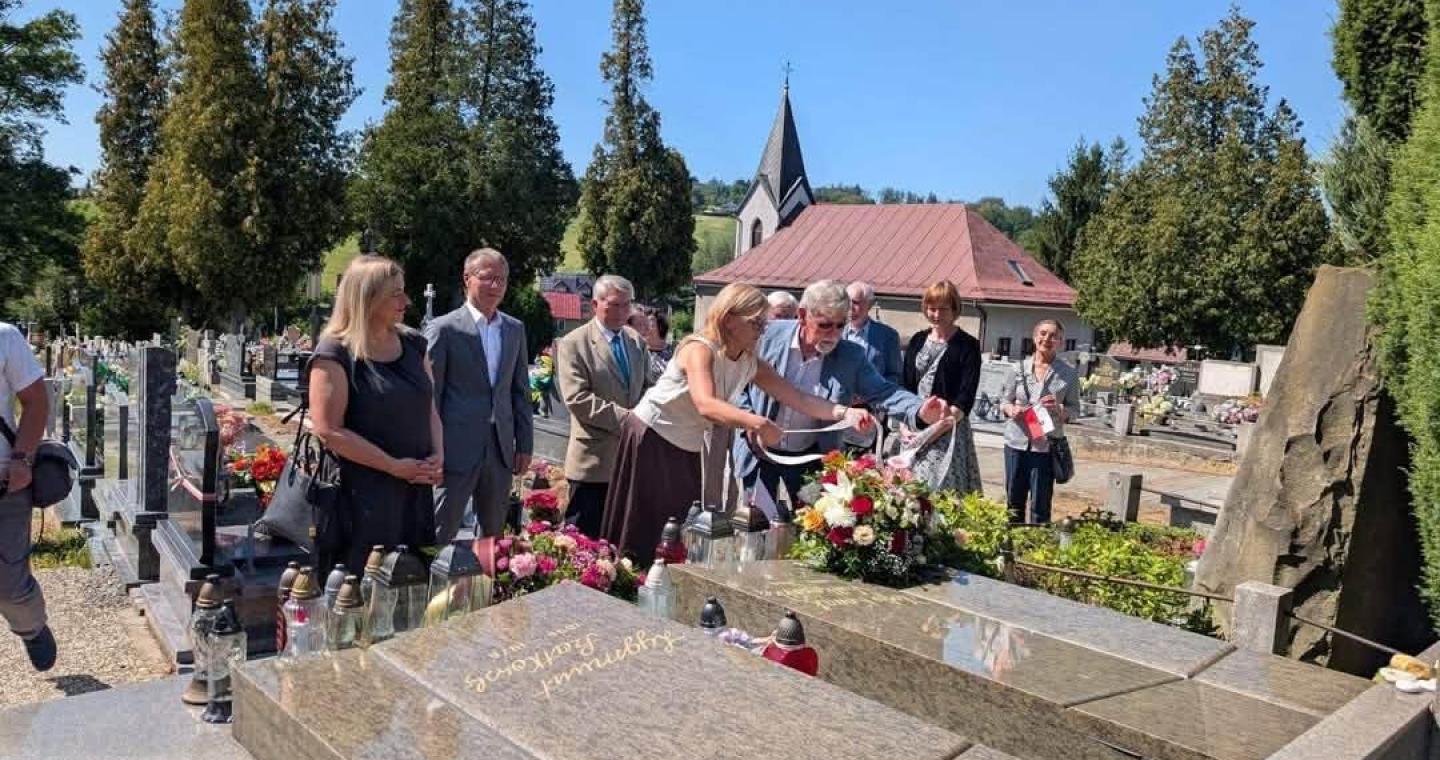 Grupa osób stoi przy grobie na cmentarzu. Dwie osoby na pierwszym planie układają kwiaty i znicze, a pozostali stoją obok. W tle widoczne są nagrobki, drzewa i budynek z wieżyczką.