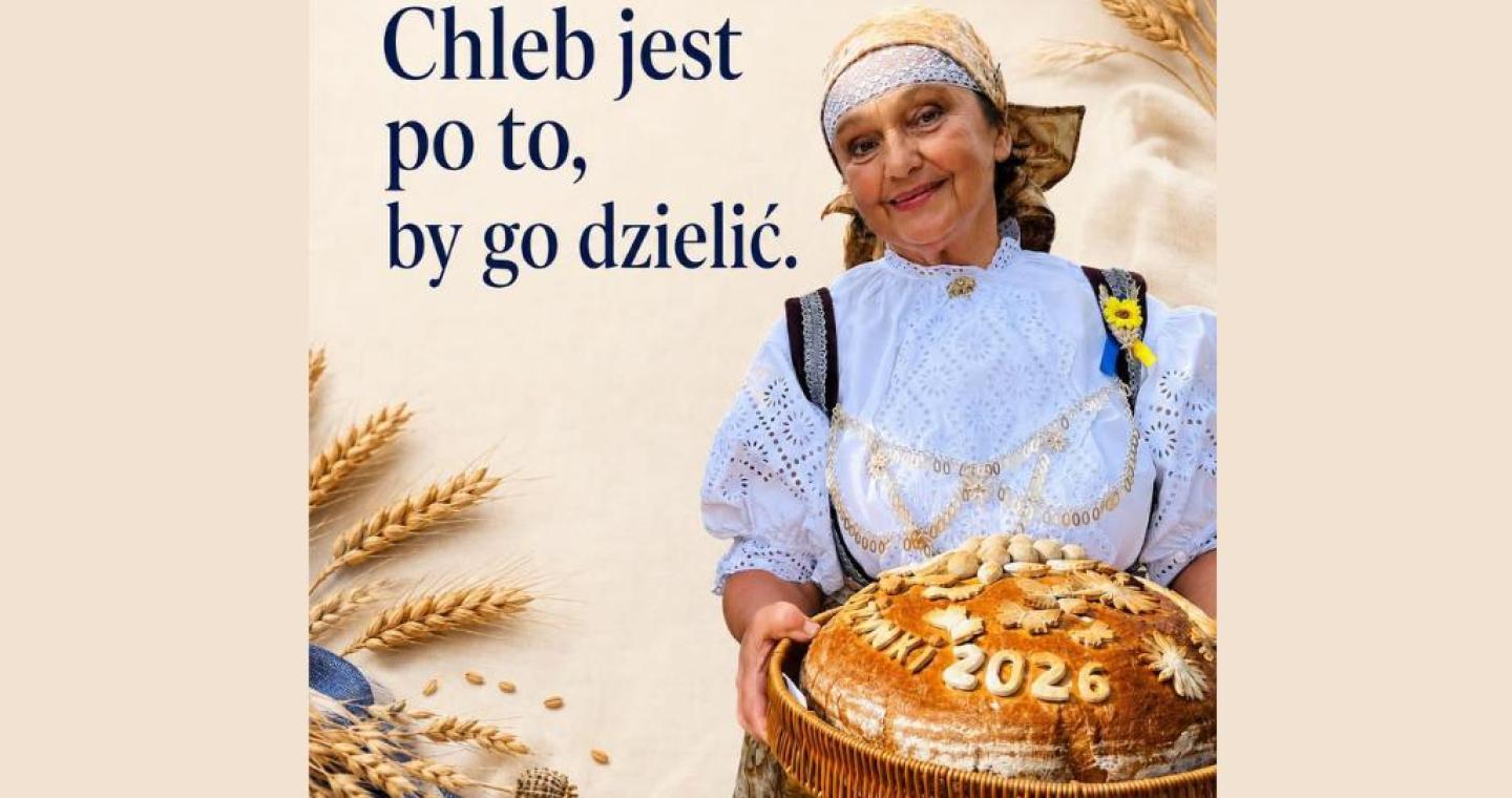 Beżowa grafika dożynkowa z kłosami zbóż i napisem „Dożynki 2026. Chleb jest po to, by go dzielić.” Po prawej stronie stoi Janina Żagan Starosta Cieszyńska w stroju regionalnym, trzymająca w wiklinowym koszu duży, ozdobny bochen chleba z napisem „Dożynki 2026”.