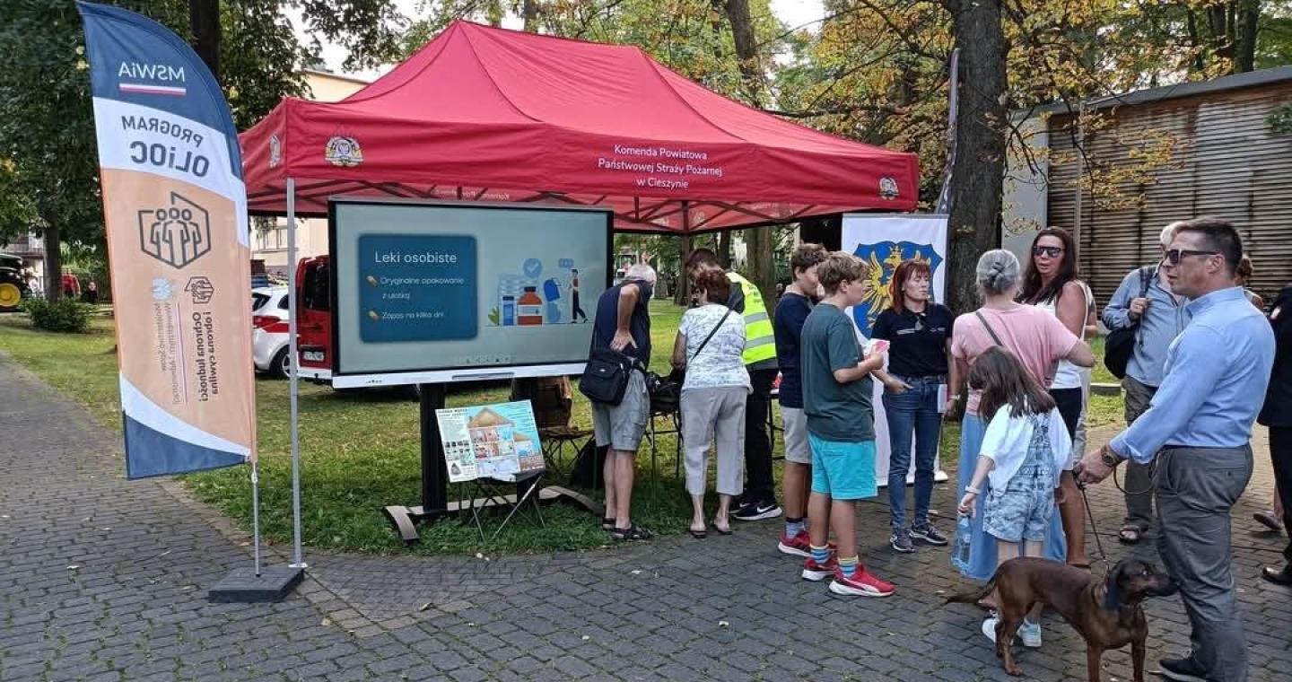 Grupa osób stoi przy czerwonym namiocie Komendy Powiatowej Państwowej Straży Pożarnej w Cieszynie . Pod namiotem znajduje się ekran z prezentacją dotyczącą leków osobistych oraz stół z materiałami informacyjnymi. Obok ustawiono flagę programu OLiOC, a na pierwszym planie stoją uczestnicy.
