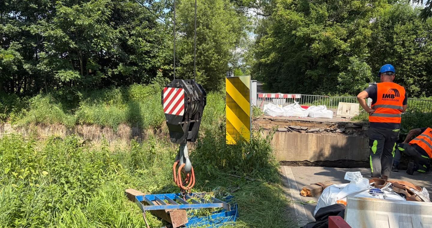 Prace przy uszkodzonym moście. Widoczny jest hak dźwigu, bariery zabezpieczające, materiały budowlane oraz pracownik w odzieży odblaskowej. Wokół znajduje się gęsta zieleń.