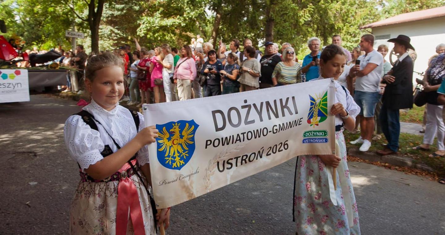 Dwie dziewczynki w strojach regionalnych niosą baner z napisem „Dożynki Powiatowo-Gminne Ustroń 2026”. W tle stoją licznie zgromadzeni uczestnicy i widzowie korowodu.