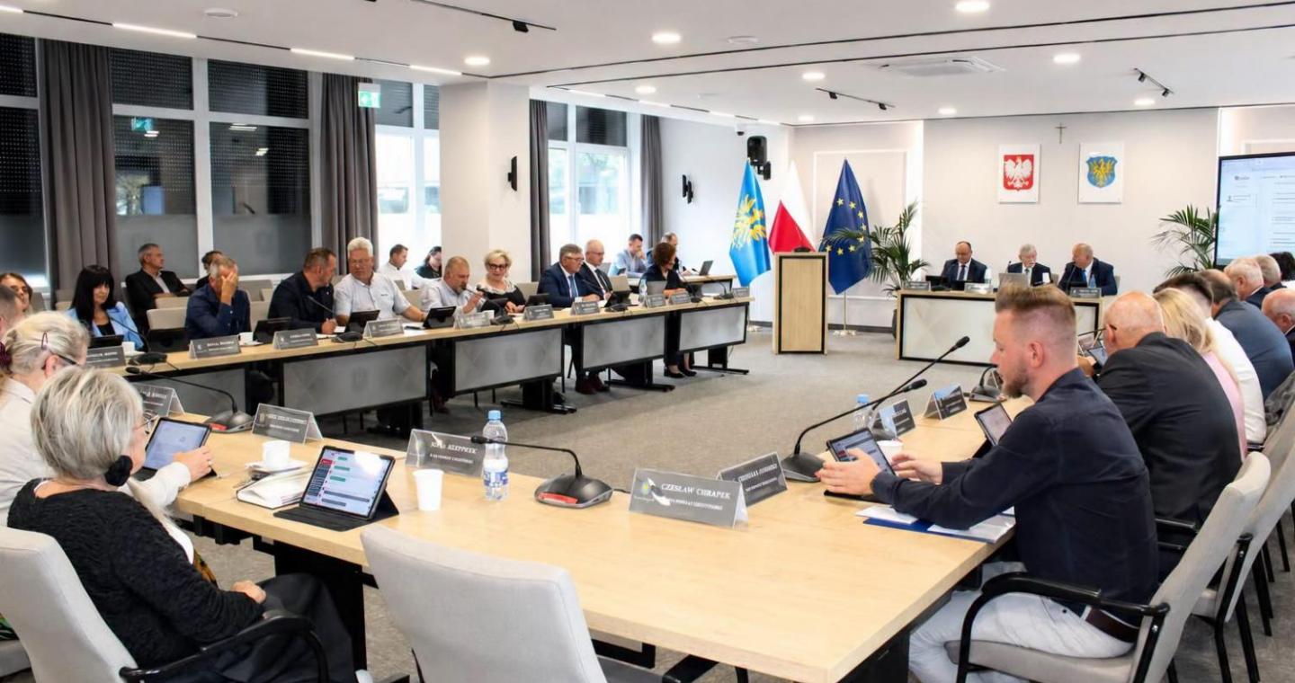 Widok na salę sesyjną podczas obrad. Uczestnicy siedzą przy ustawionych w podkowę stołach, przed nimi znajdują się mikrofony, tablety i tabliczki z nazwiskami. W centralnej części sali widoczne są mównica oraz flagi, a na ścianie godło Polski i herb Powiatu Cieszyńskiego.