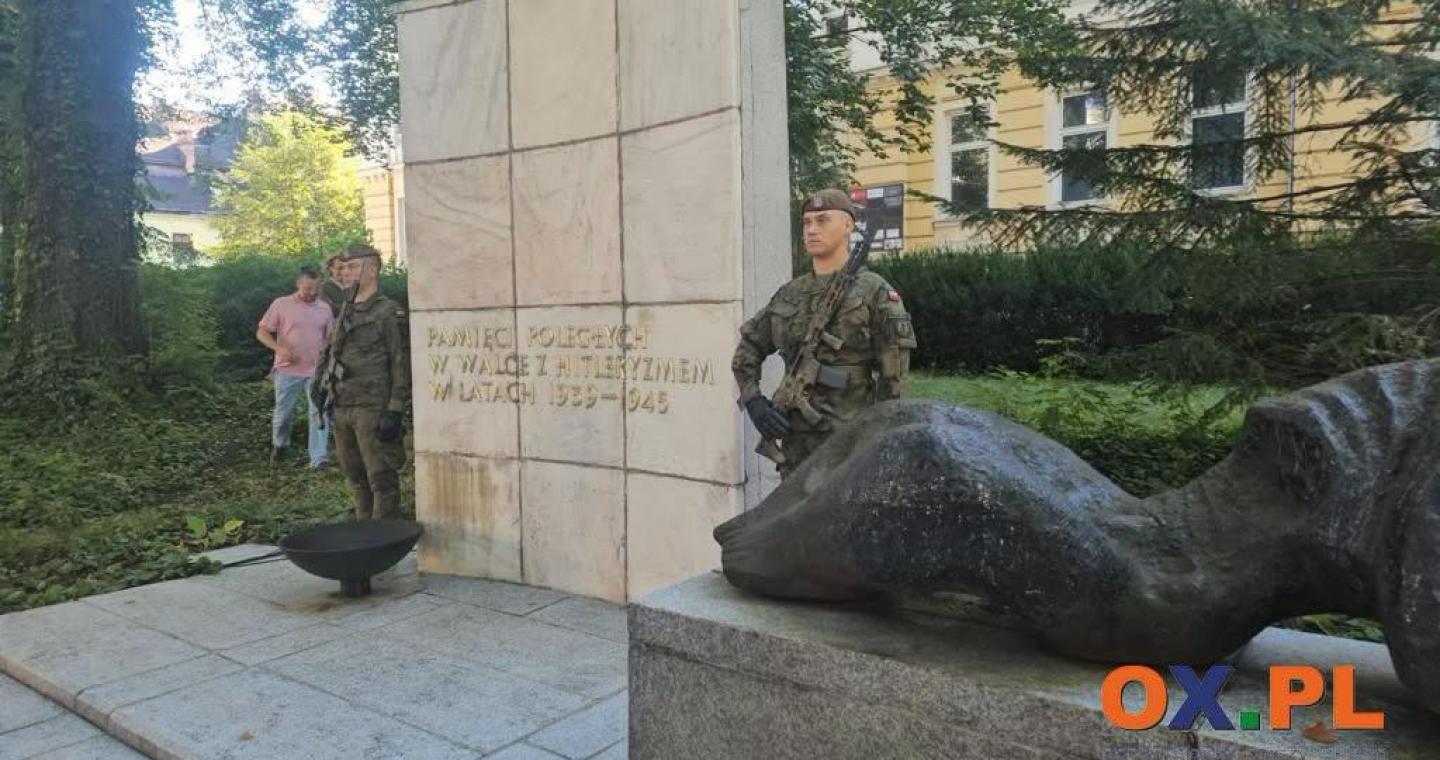 Pomnik z jasną kamienną płytą z napisem oraz ciemną rzeźbą umieszczoną na cokole. Po obu stronach monumentu stoją żołnierze w mundurach, a w tle widoczne są drzewa, krzewy i budynek.